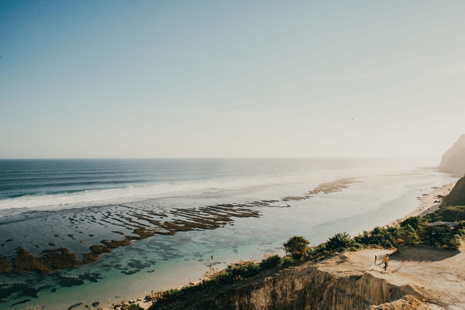 A couple walking on a rocky beach below a green cliff on the Bali coast.